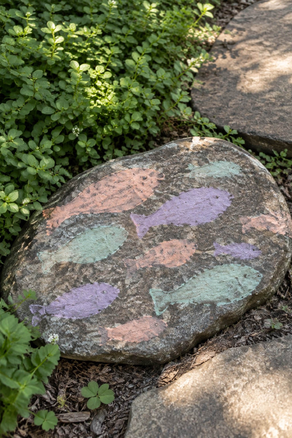 Large rock with colorful painted fish in orange, purple, and teal amid green plants.