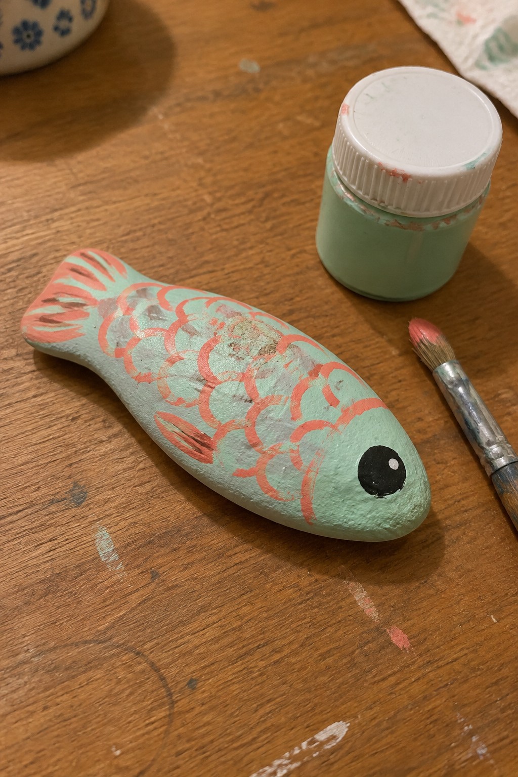 Mint green painted wooden fish with coral scales beside paint jar and brush.