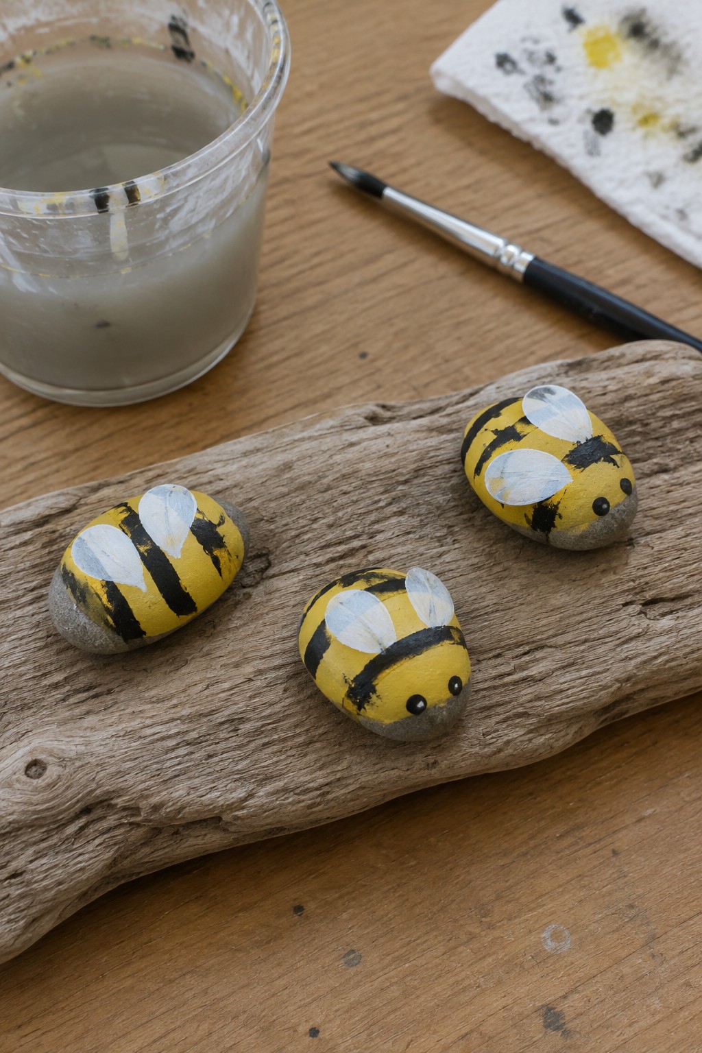 Three yellow-and-black bee rocks on driftwood beside paint jar and brush.