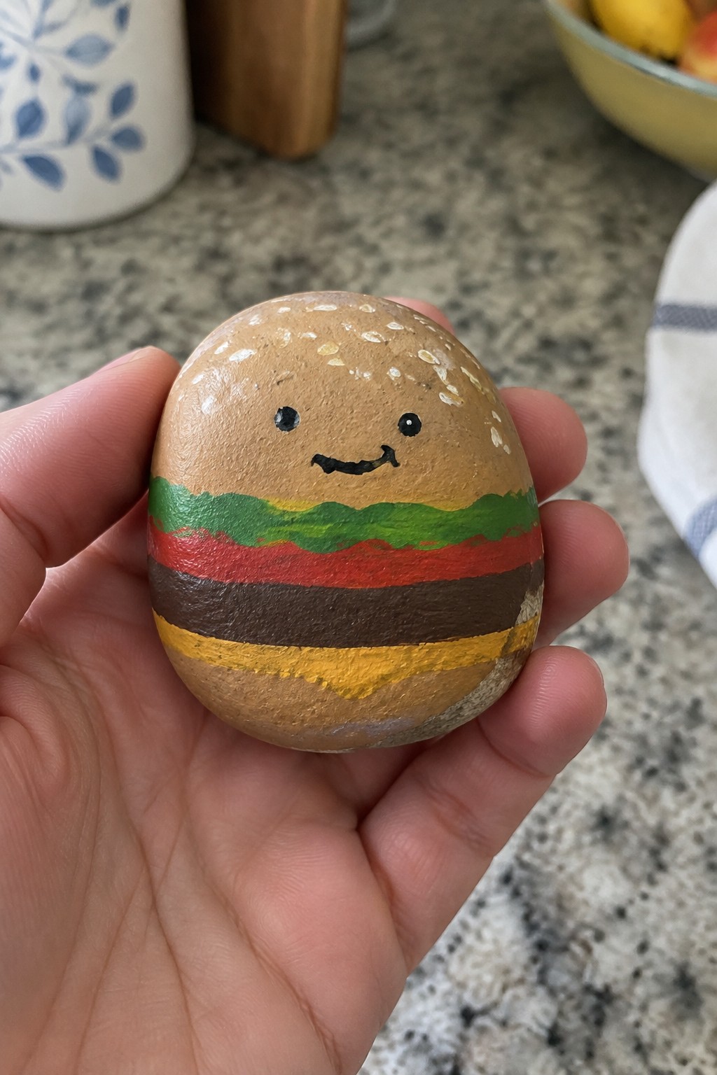 Hand holding a smiling hamburger painted on a round stone with colorful layers.