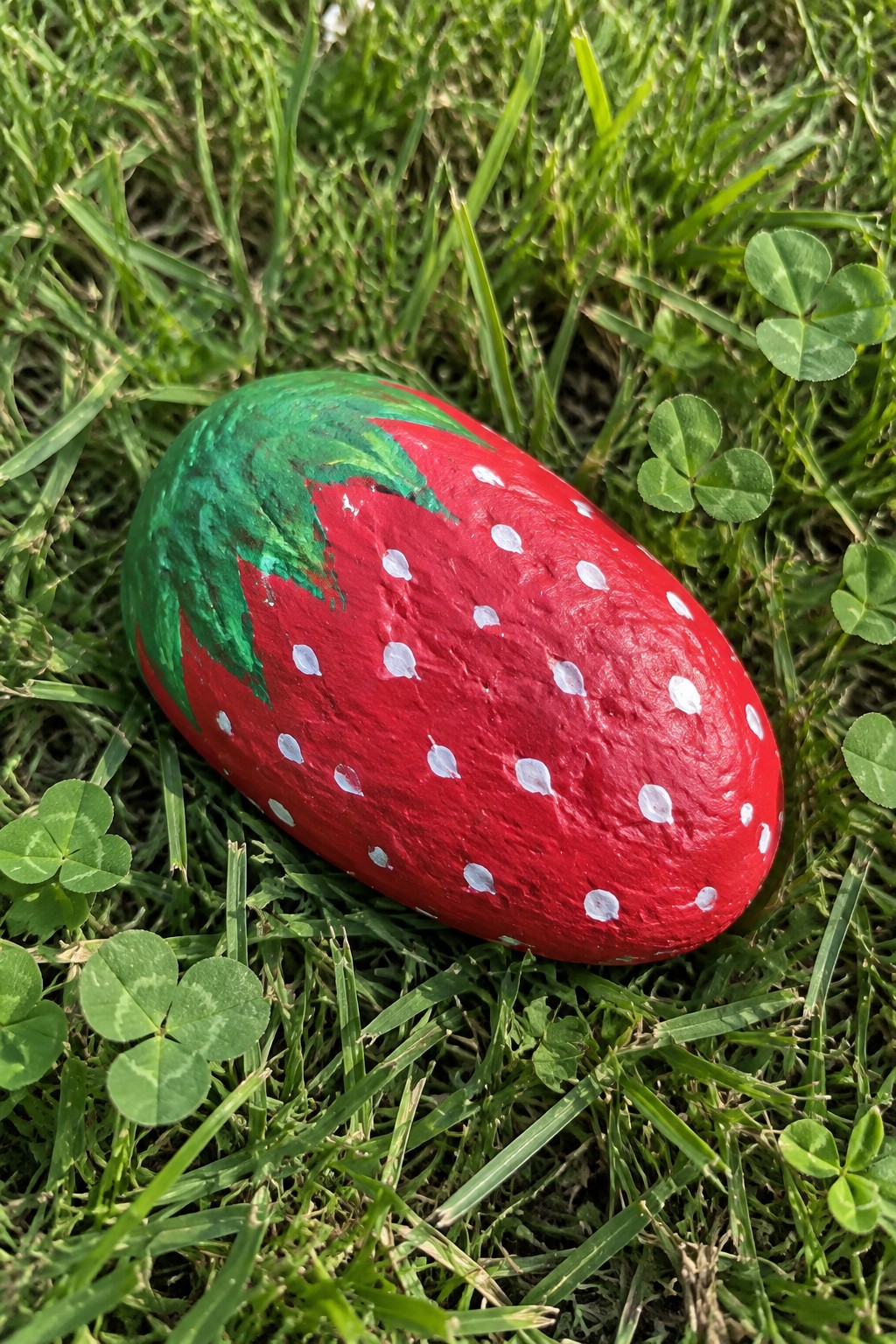 Red strawberry-painted rock with white dots and green leaves on grassy clover