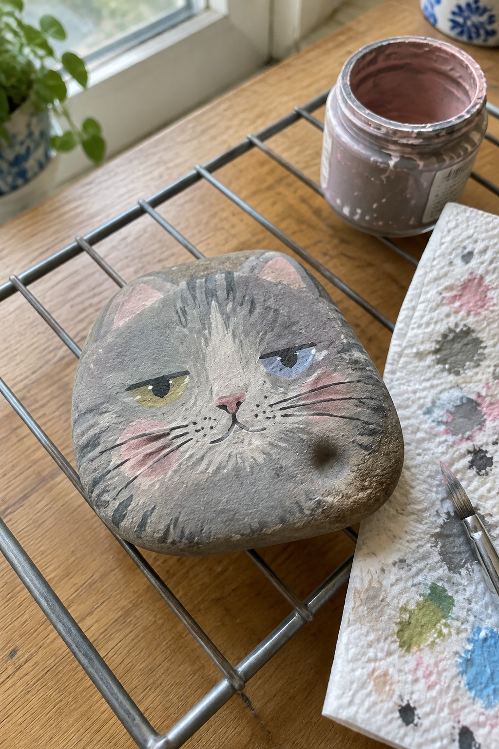 Grumpy gray cat face painted on stone atop metal rack beside paint jar