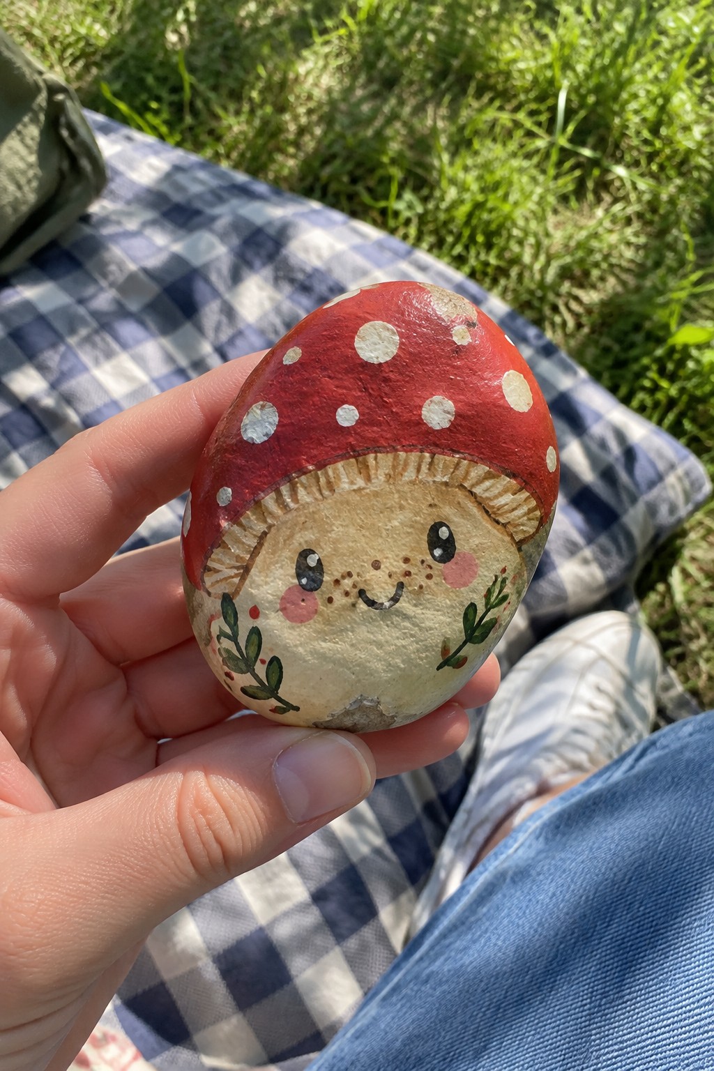 Hand holding a red mushroom rock with white spots and smiling face on checkered blanket.