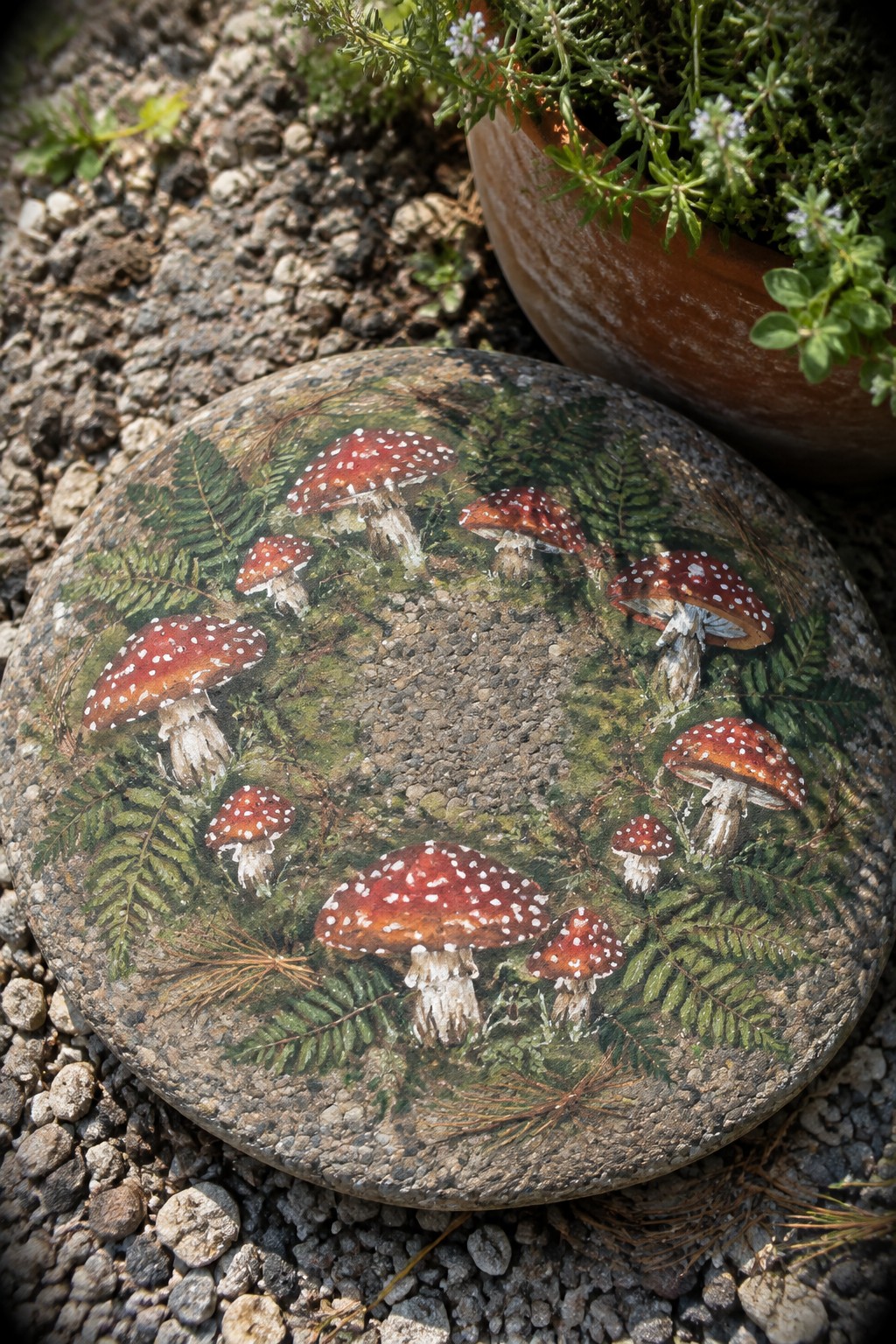 Round stone painted with red white-spotted mushrooms amid green ferns on gravel