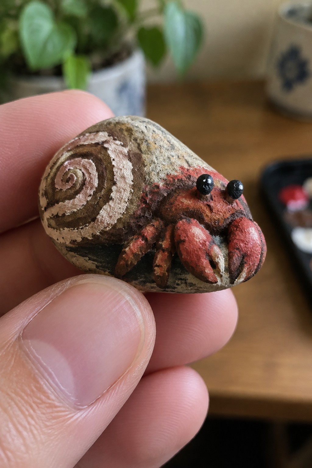 A small painted rock held in a hand shows a hermit crab design with a brown spiral shell pattern on the back and red claws and body in front.