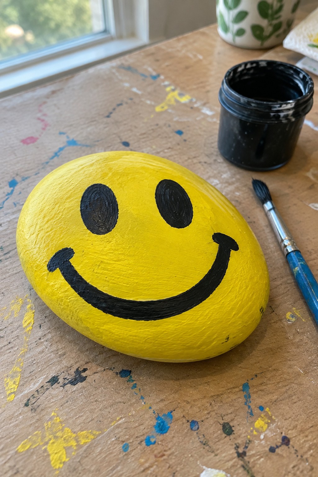 Yellow smiley-face rock on paint-splattered table beside black paint jar and brush
