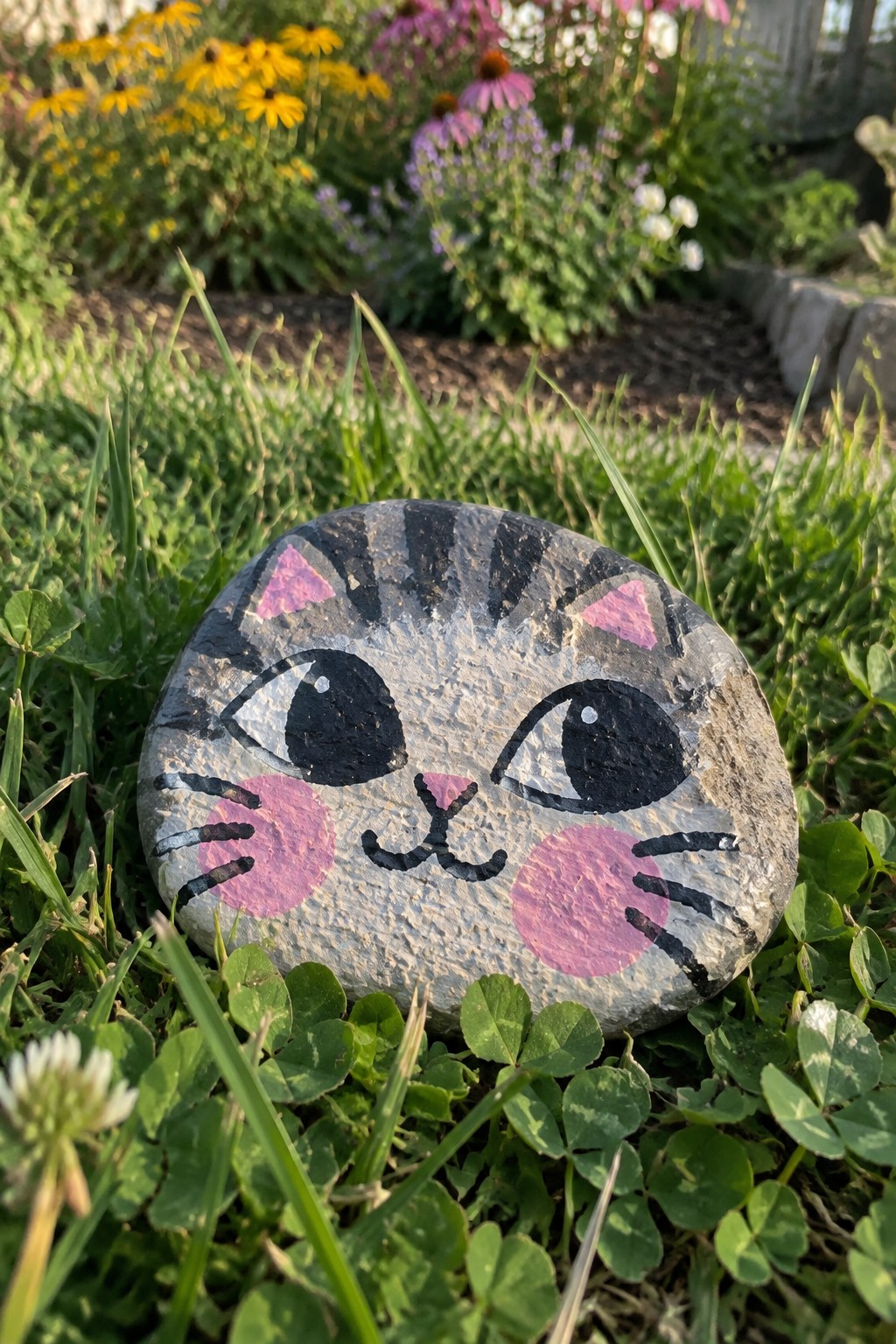 Painted cat-face rock with pink cheeks on green grass amid garden flowers