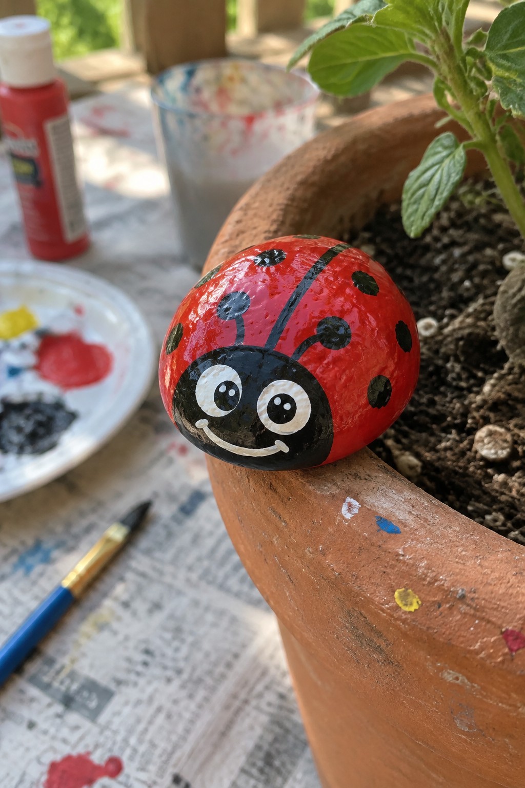 Smiling ladybug rock on terracotta pot rim amid paint supplies and brush.