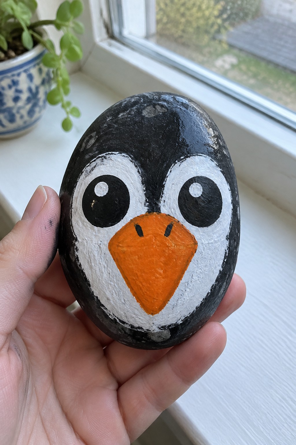Hand holding black rock painted as penguin with large eyes and orange beak