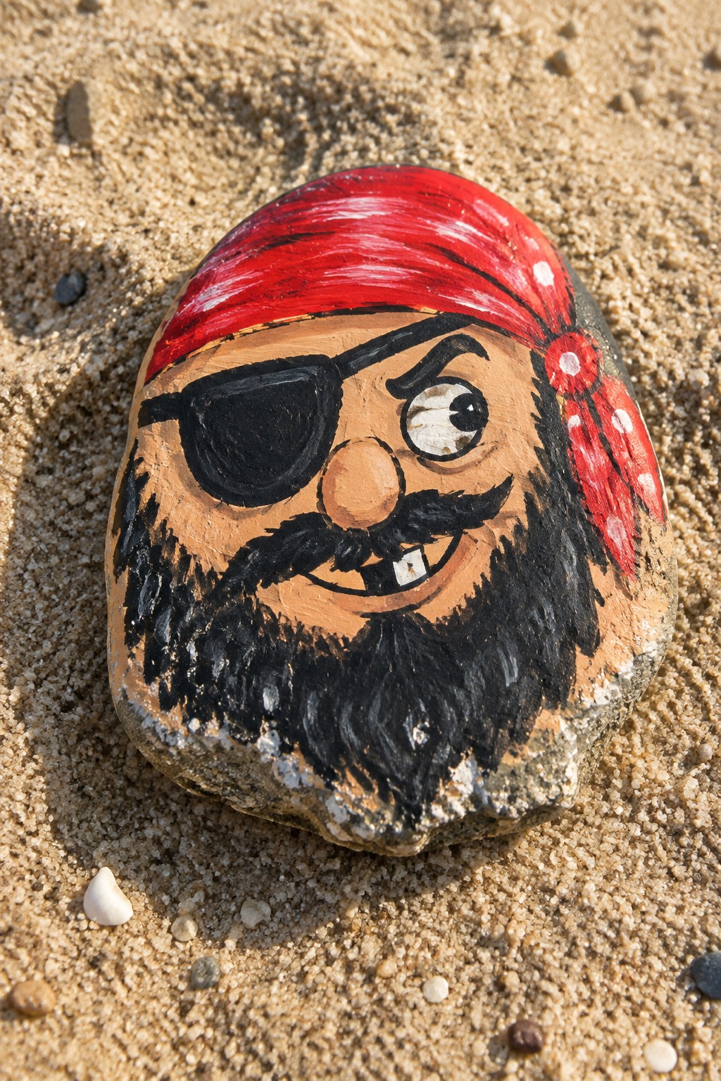 Painted rock on sand showing pirate face with red bandana, eyepatch, and black beard.