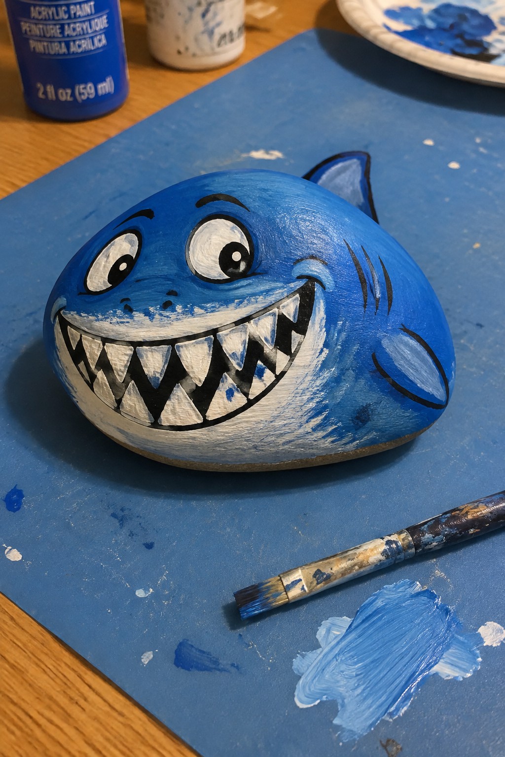 Blue painted shark rock with cartoon eyes and teeth beside paintbrush on blue mat