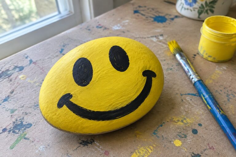 20 Painted Rocks for Kids With Bold Cartoon Faces Yellow smiley-face rock on paint-splattered table beside black paint jar and brush