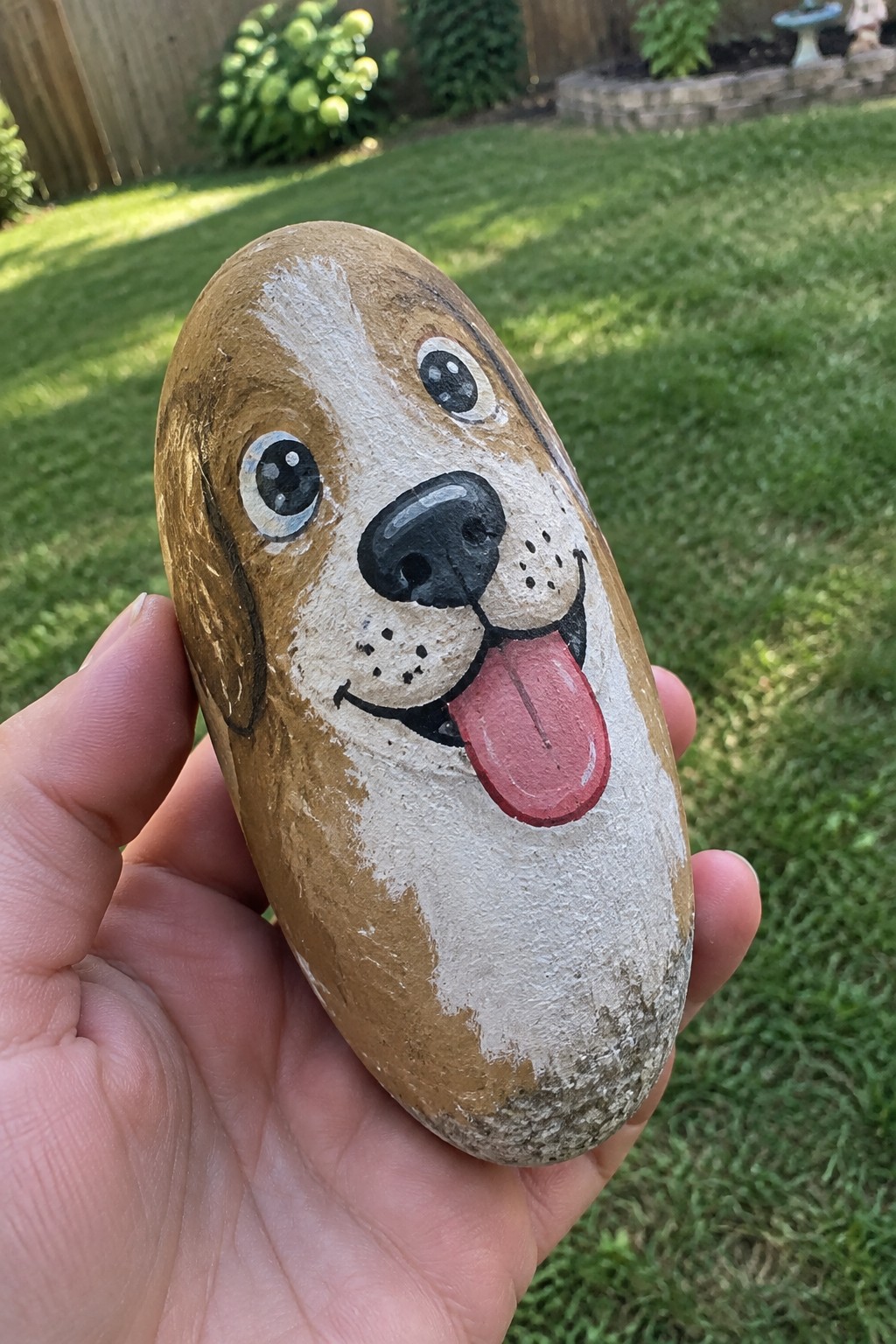 Hand holding oval rock painted as brown-and-white dog with pink tongue out