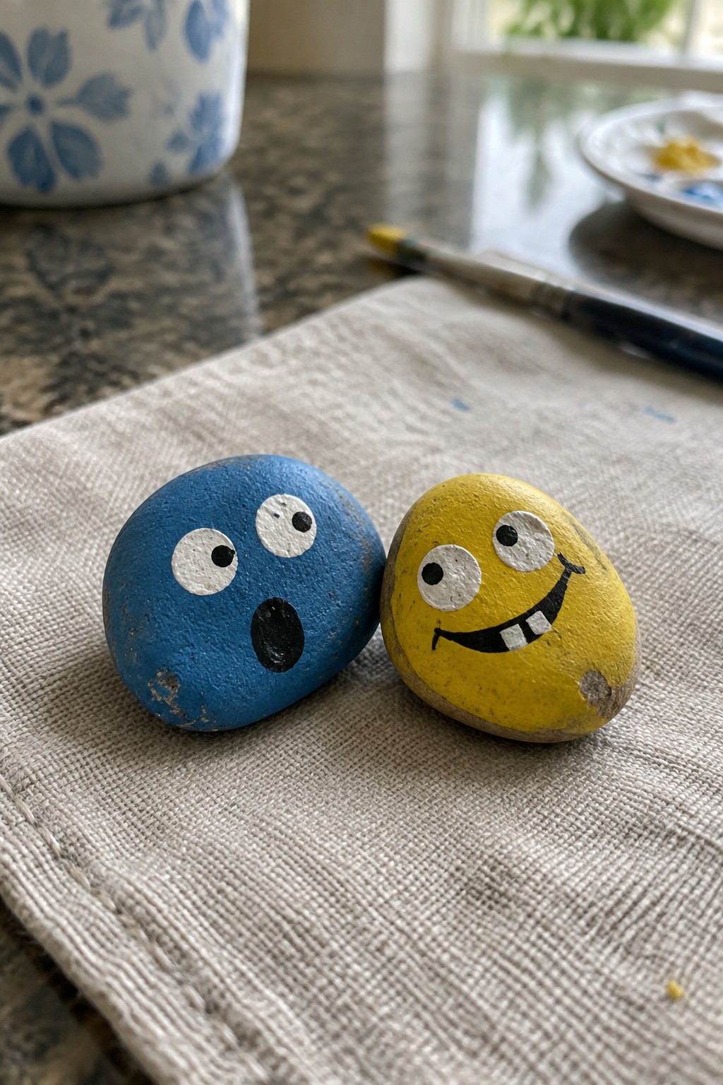Blue surprised and yellow grinning painted rocks on beige cloth with paintbrush nearby
