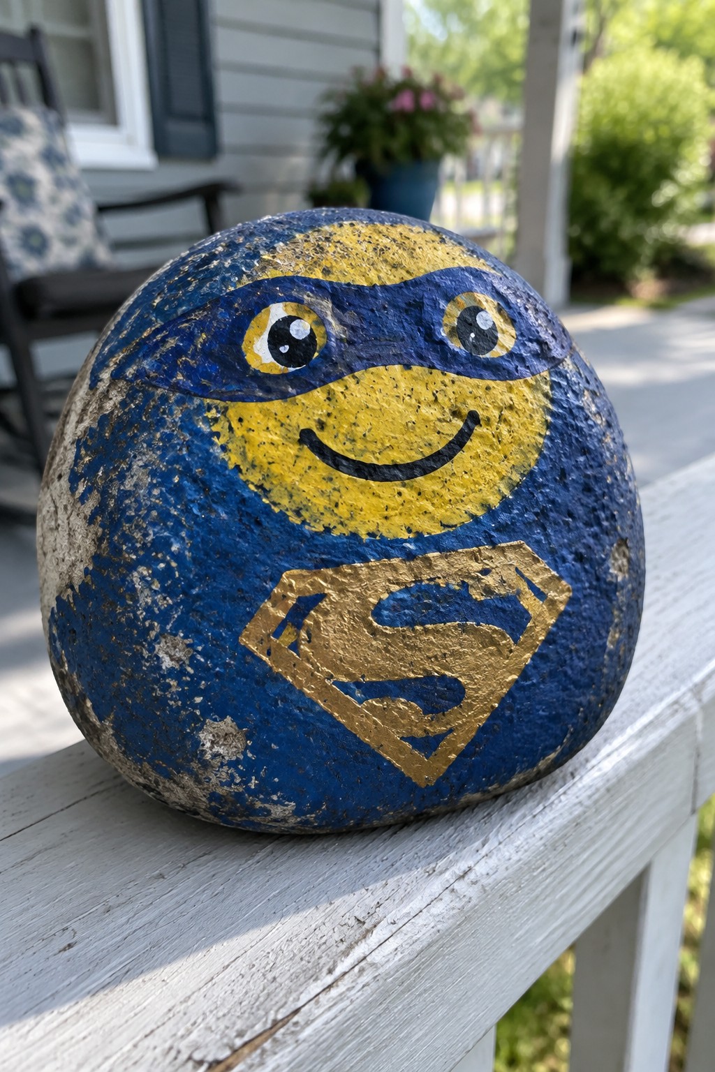 Blue painted rock with smiling yellow face, blue mask, and gold Superman logo on railing