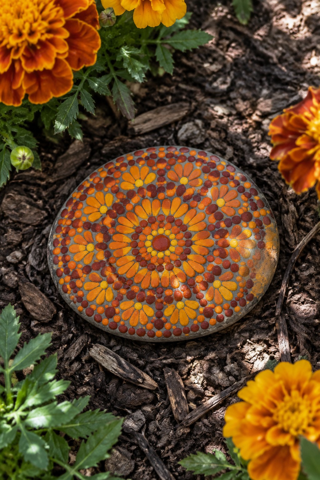 Colorful painted stone with orange floral mandala pattern amid blooming marigolds