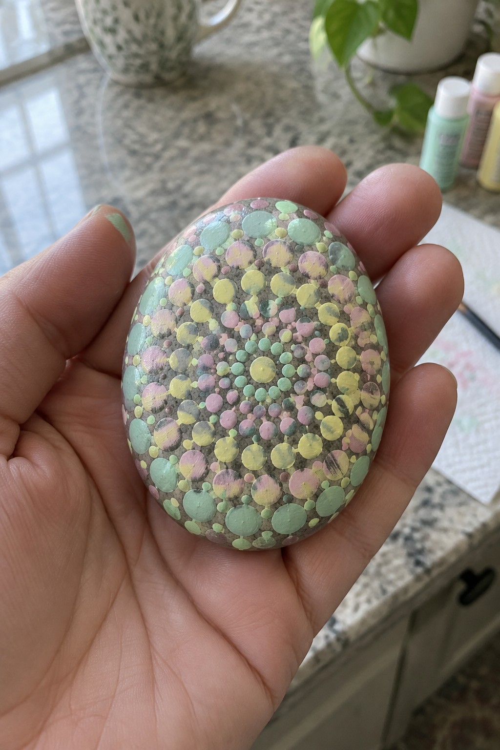 Hand holding oval stone painted with pastel green, yellow, and pink dots