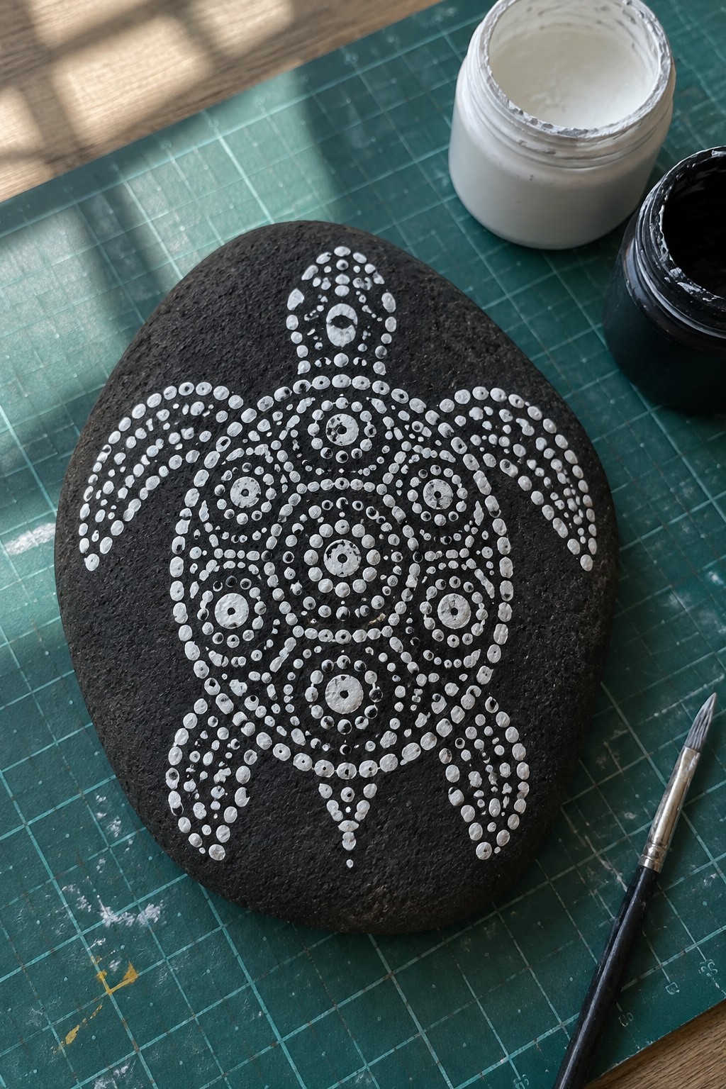 Black stone with white dotted turtle on green mat beside paint jars and brush