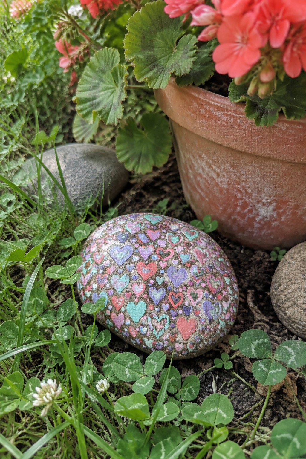 Painted rock covered in colorful hearts amid clover and potted pink flowers