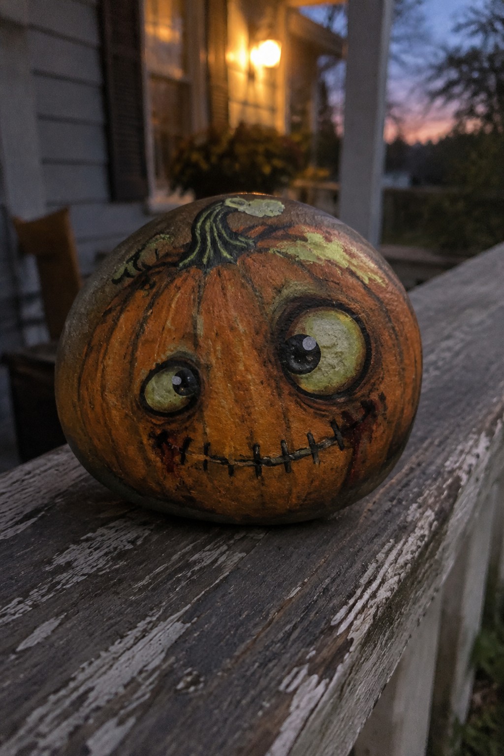 Painted pumpkin with mismatched eyes and stitched mouth on weathered wooden railing at dusk