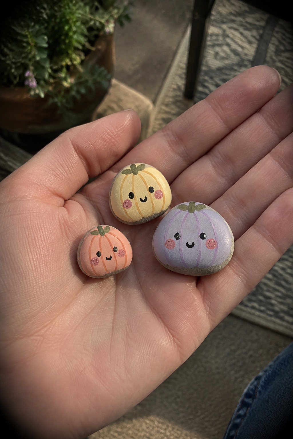 Hand holding three small painted pumpkin rocks with cute faces in pastel colors.