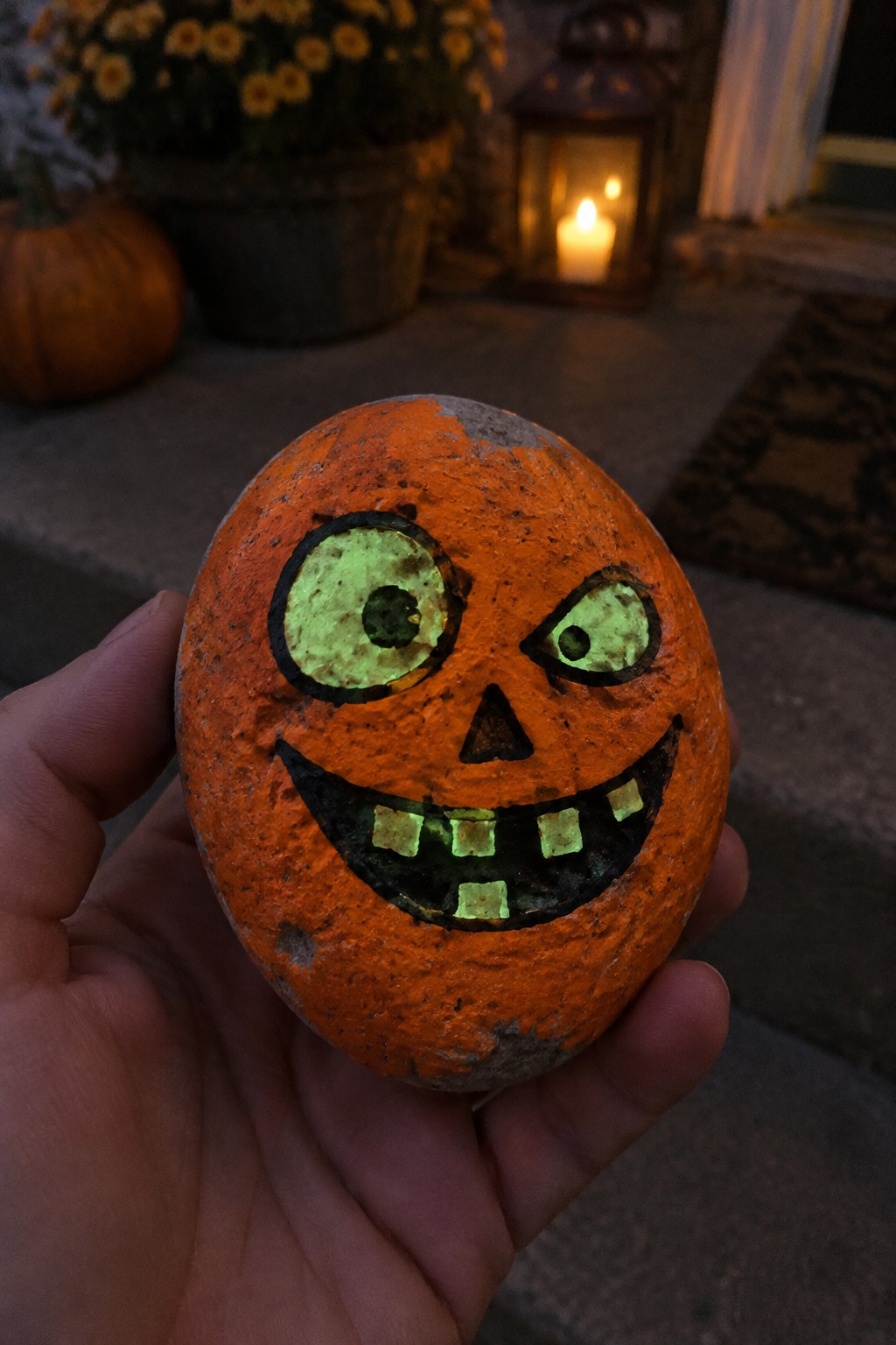 Hand holding orange rock painted as glowing green jack-o'-lantern face