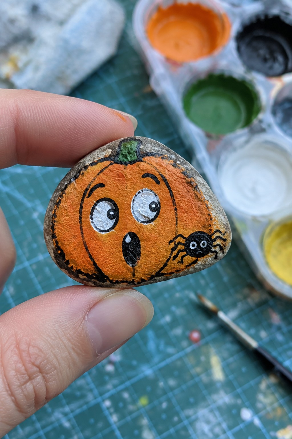 Hand holding painted rock of surprised orange pumpkin with spider
