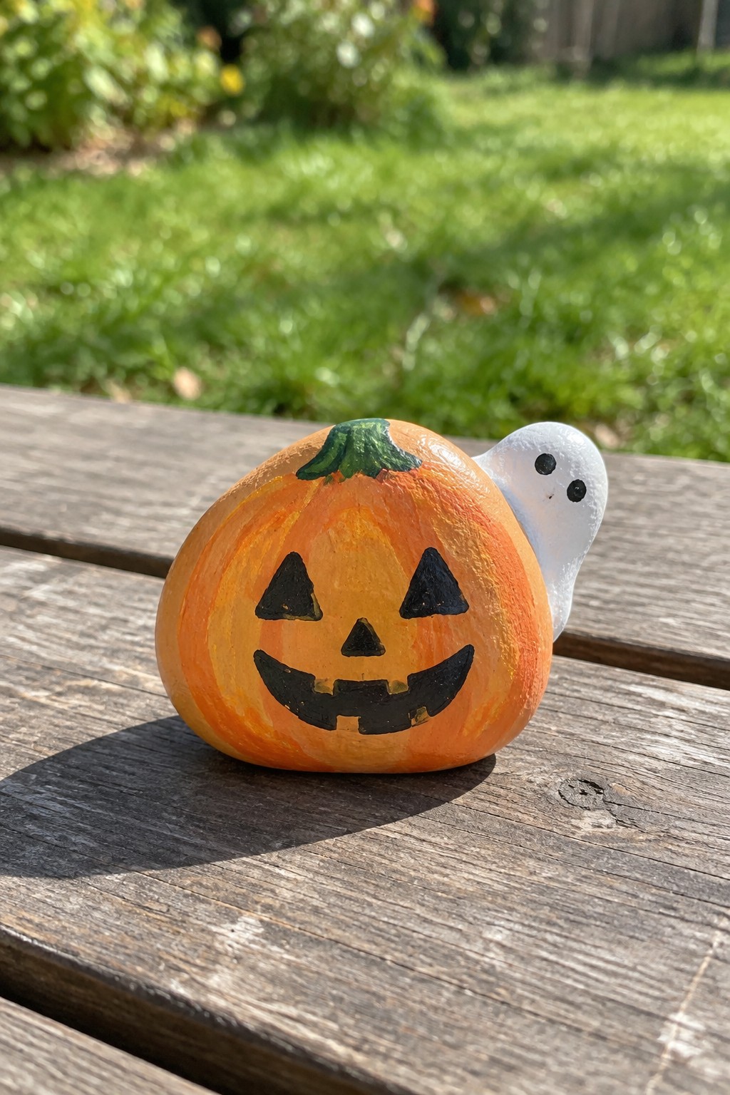 A painted orange pumpkin rock with a jack-o-lantern face and a small white ghost on the side sits on a wooden outdoor surface.
