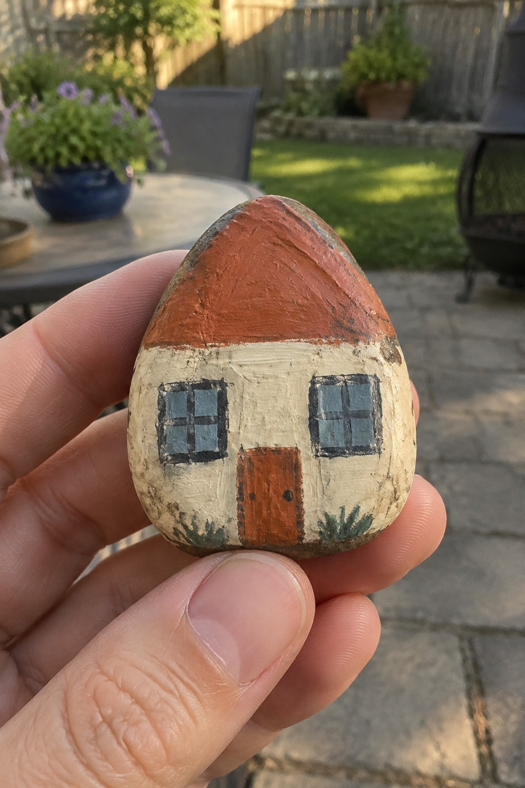 Hand holding egg-shaped rock painted as house with red roof and blue windows