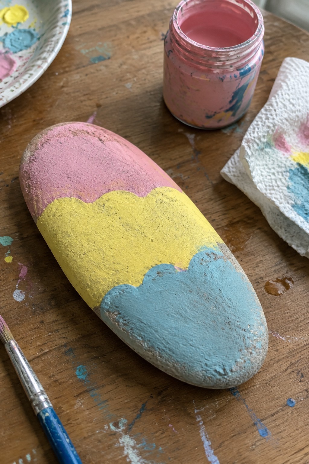 An oval rock painted with pink, yellow, and blue sections separated by scalloped lines rests on a wooden table beside paint jars and a brush.