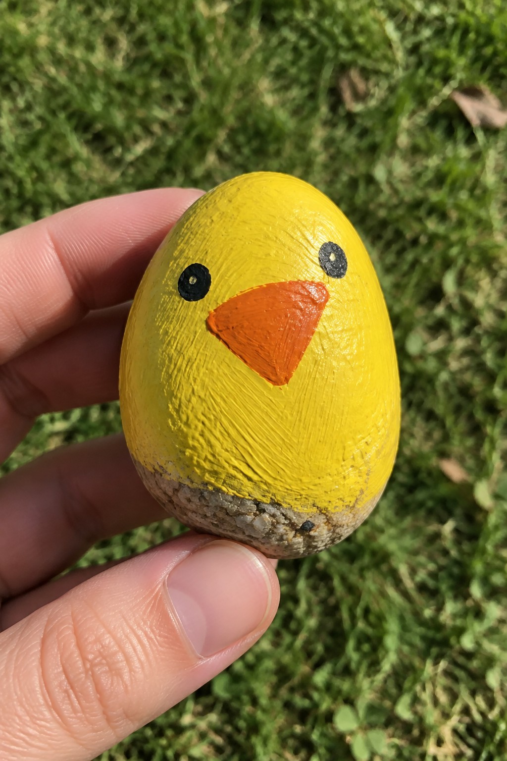 Hand holding yellow painted rock chick with orange beak and black eyes on grass