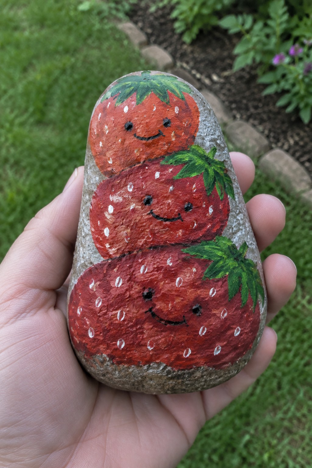 Hand holding rock painted as three stacked smiling red strawberries with green leaves