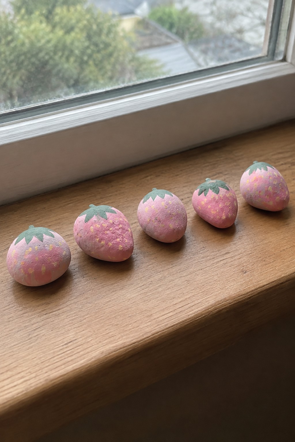 Five pink strawberry rocks with green tops on a wooden windowsill.
