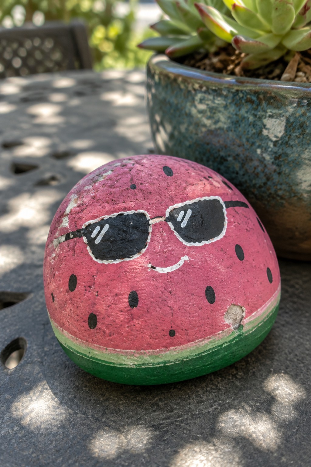 Painted watermelon rock with sunglasses and smile on dark table near potted plant