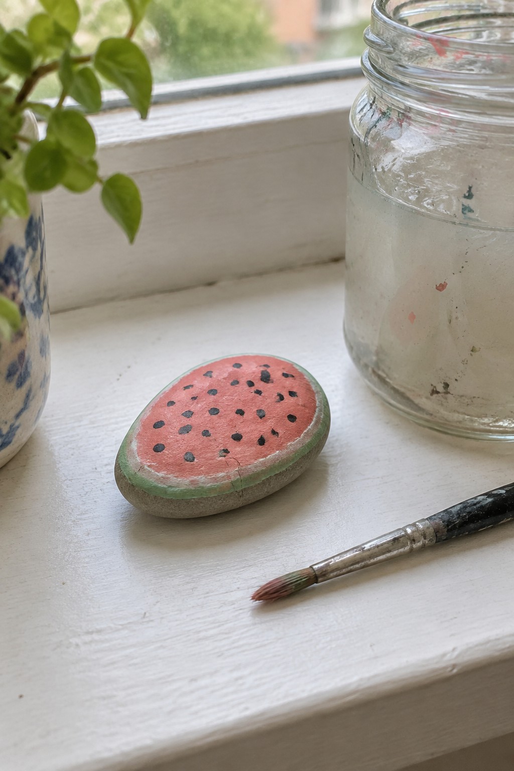 Watermelon-painted stone, paintbrush, and jar on white windowsill beside plant