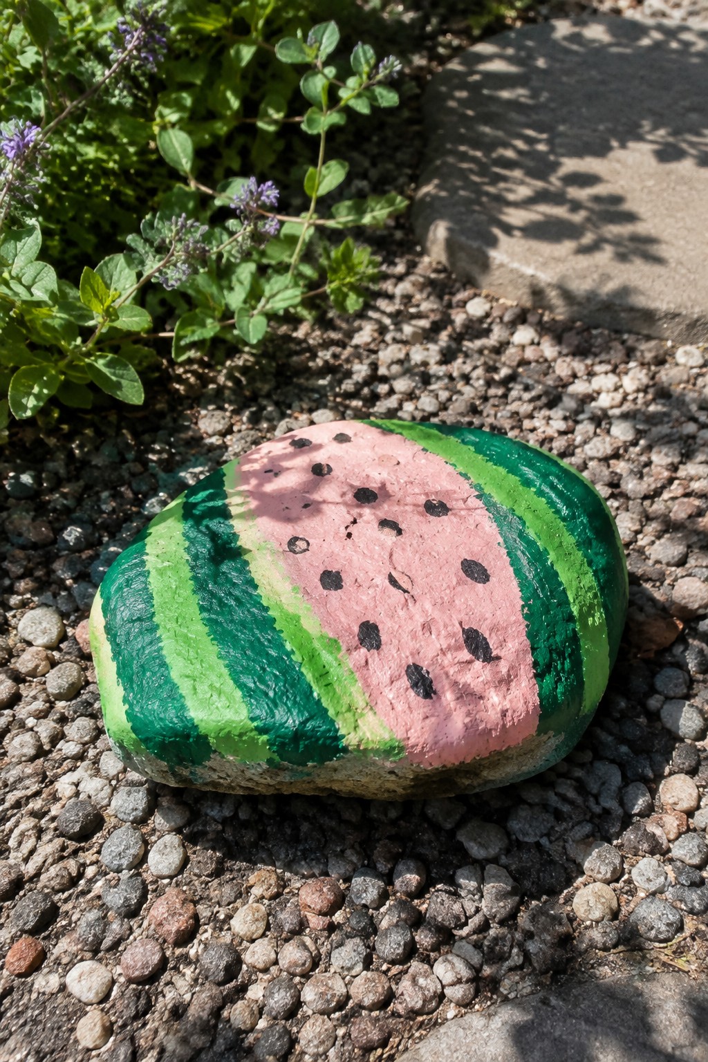 Watermelon-painted rock with green stripes, pink center, and black seeds on gravel.