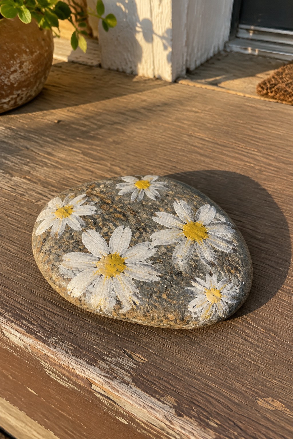 Painted stone with white and yellow daisies on sunlit wooden surface