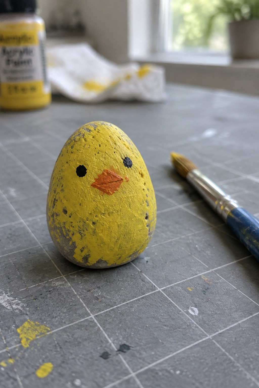 Yellow painted rock chick with black eyes and orange beak beside paintbrush