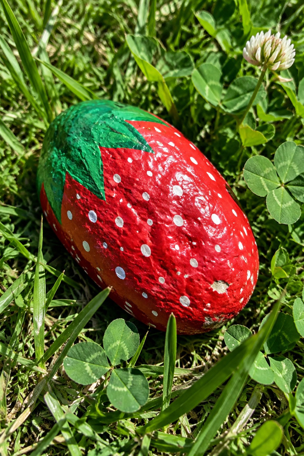 Red strawberry-painted rock with white dots and green leaves on grass