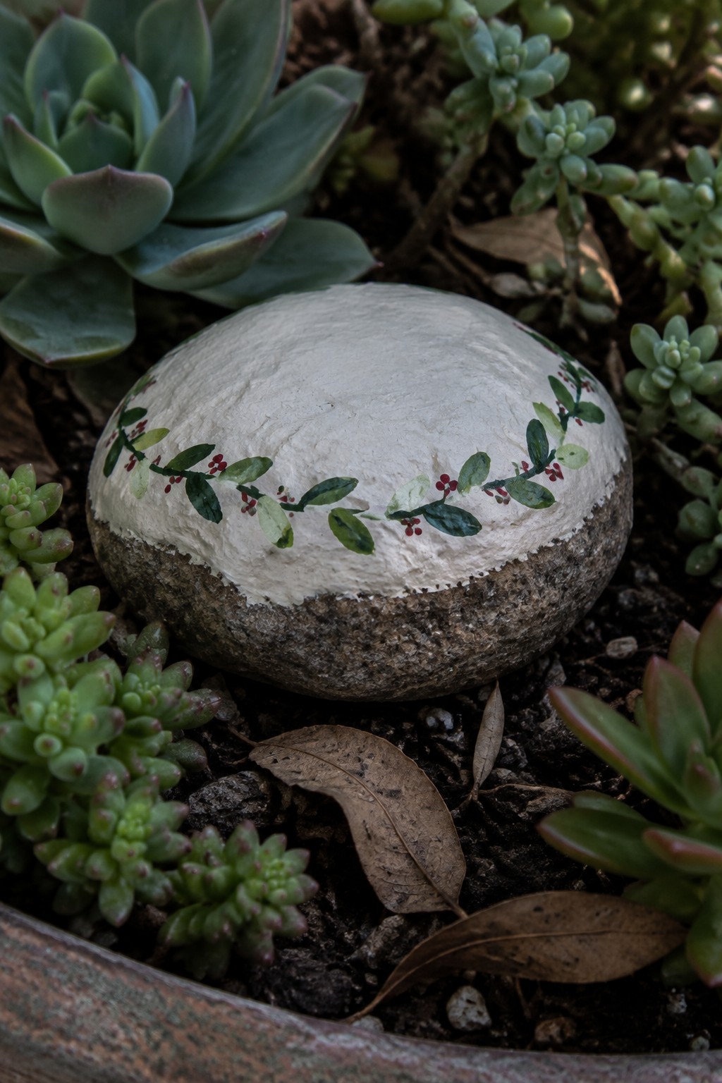 Painted white rock with green vine and red berries among succulents and dry leaves