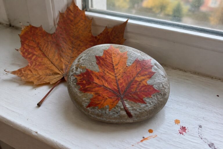 20 Fall Rock Painting Ideas With Autumn Leaves Stone with painted red-orange maple leaf beside real dried leaf on white windowsill