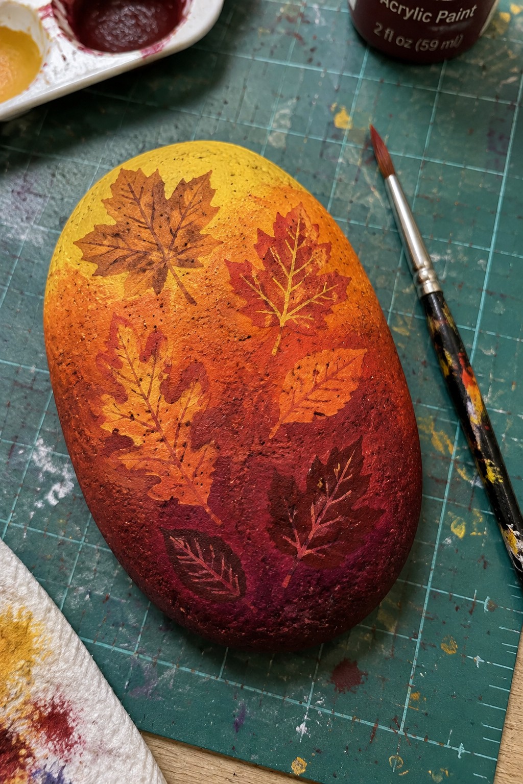 An oval painted rock shows a yellow-to-purple gradient with several autumn leaves in fall colors, sitting on a green cutting mat beside a paintbrush and palette.