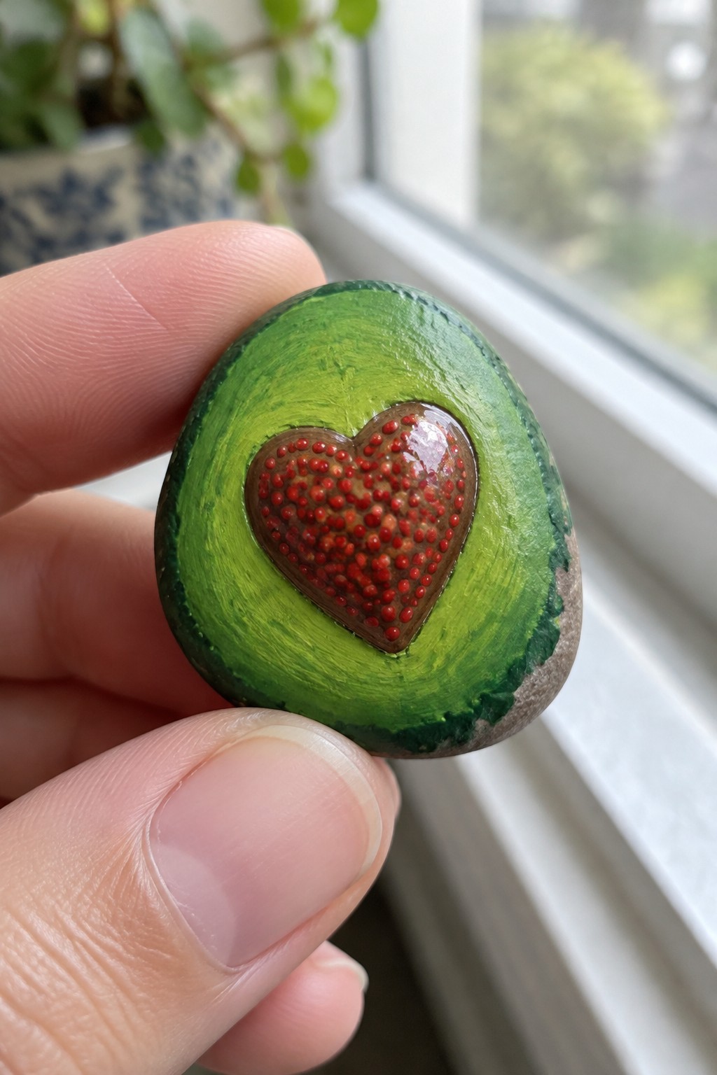 Hand holding green painted stone with glossy red-dotted heart center