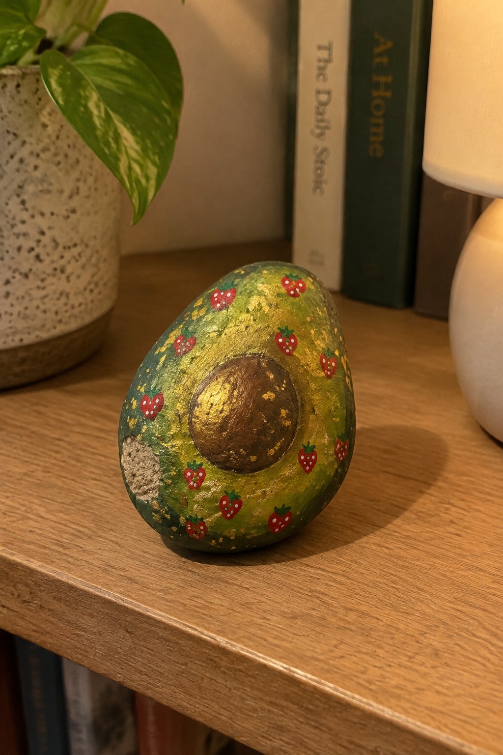 Painted avocado-shaped stone with strawberries and gold on wooden shelf near books and plant