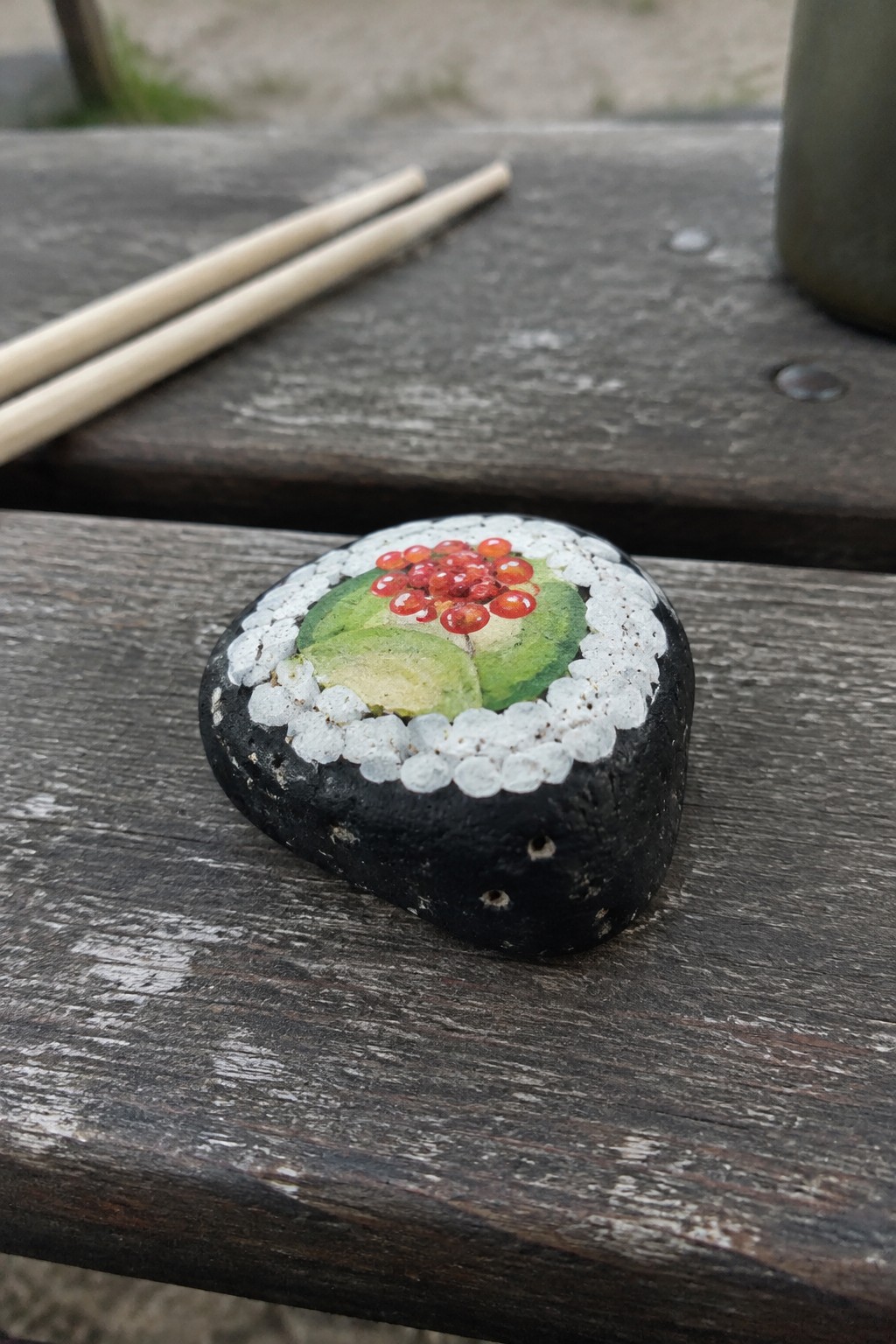 Black rock painted as sushi roll with avocado and red roe on wooden table near chopsticks