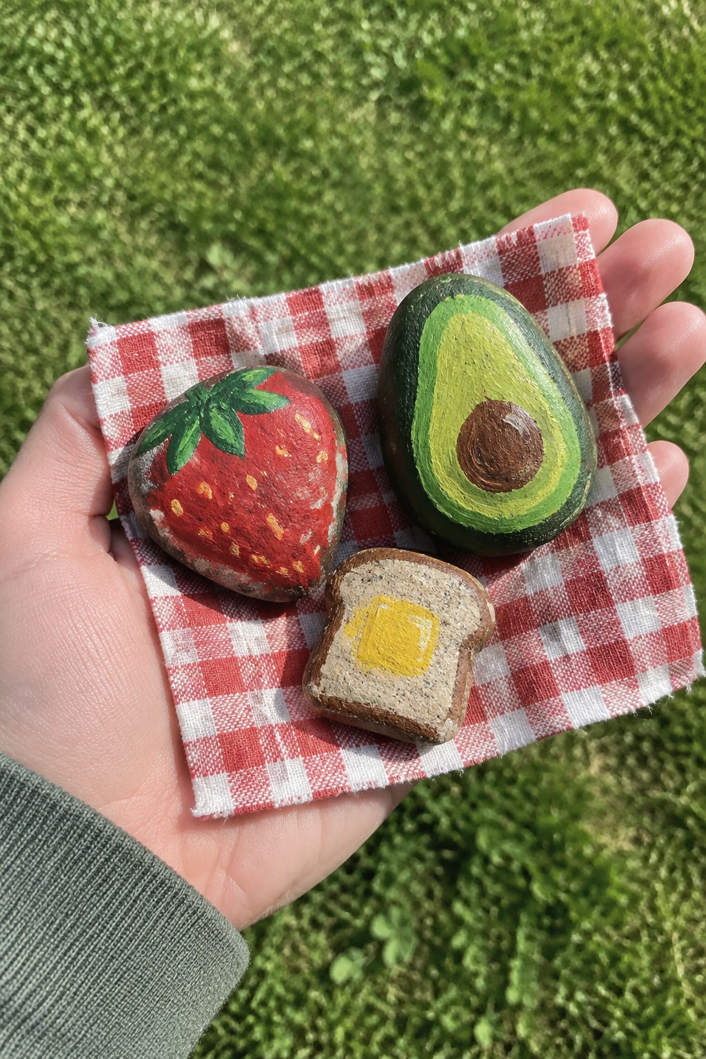 Hand holding red checkered cloth with painted strawberry, avocado, and toast rocks on grass.