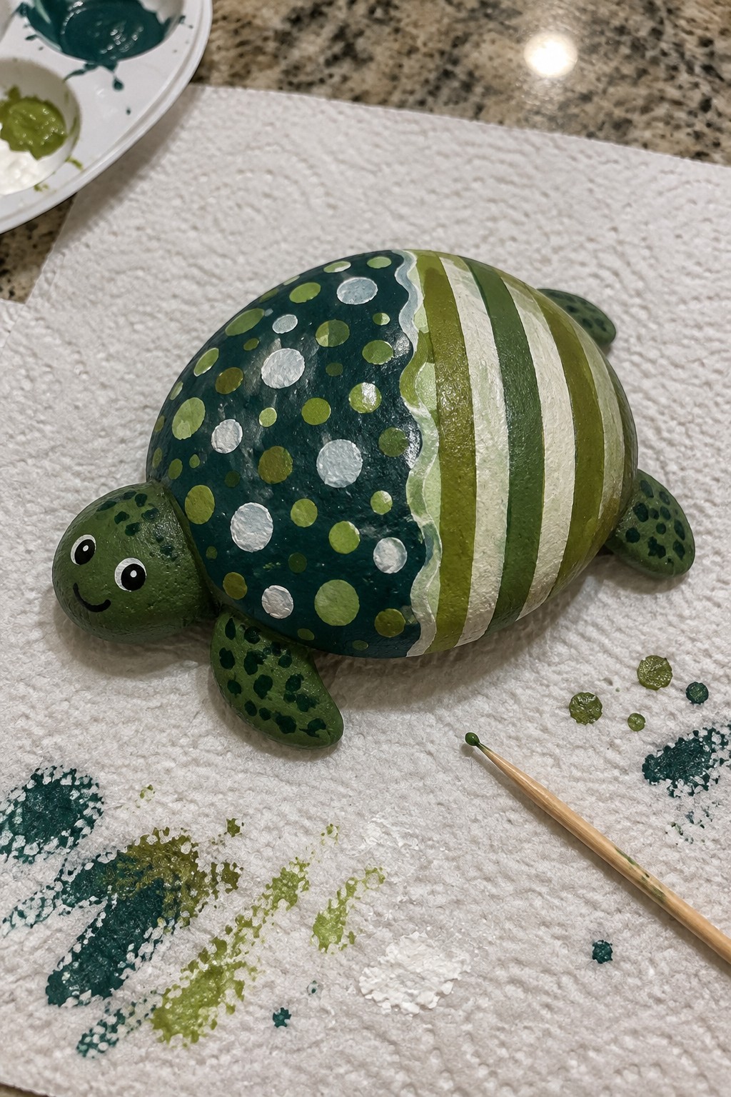 Green painted rock turtle with polka-dot shell on paper towel near paints