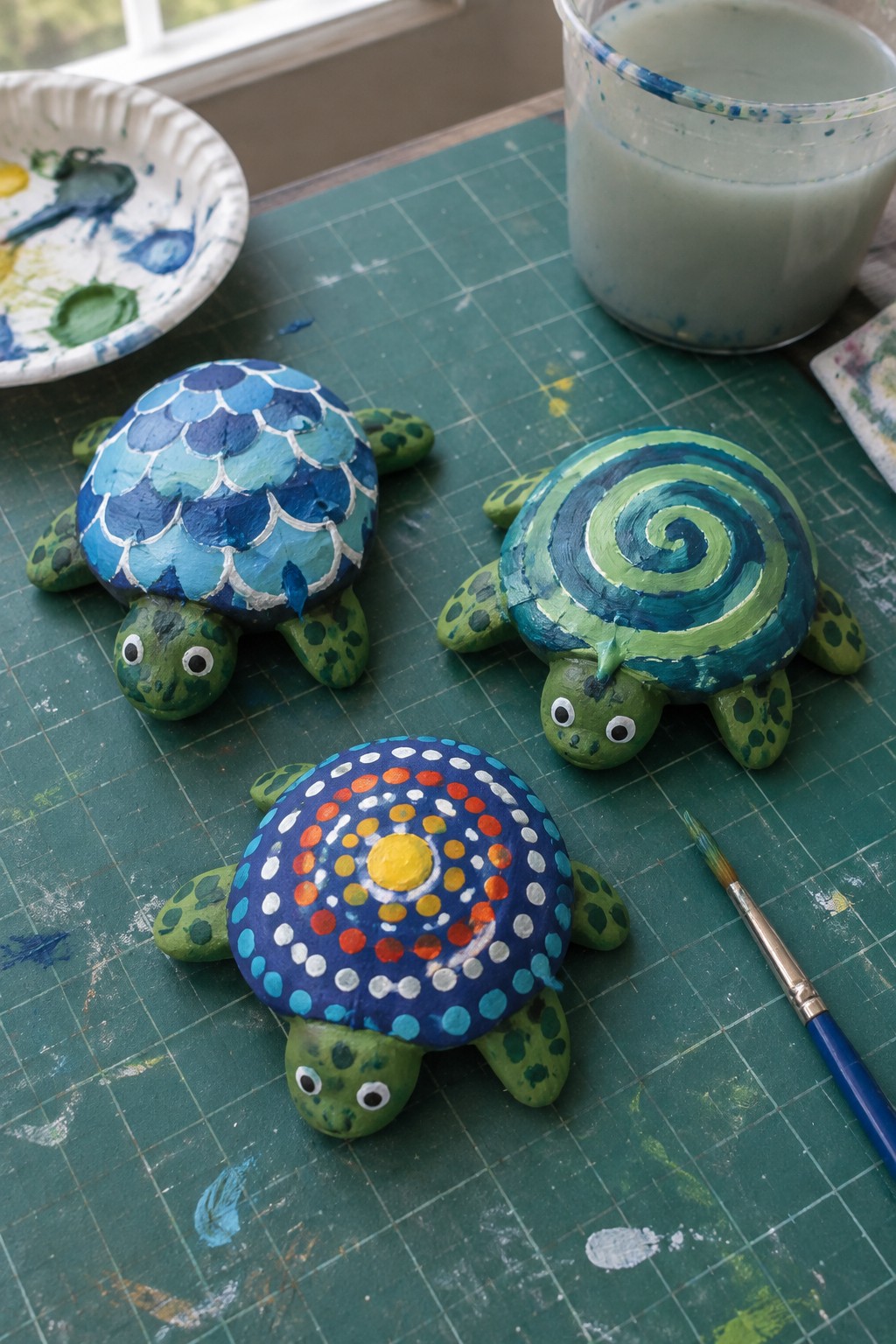 Three colorful painted turtle figurines on green craft mat with paint supplies.