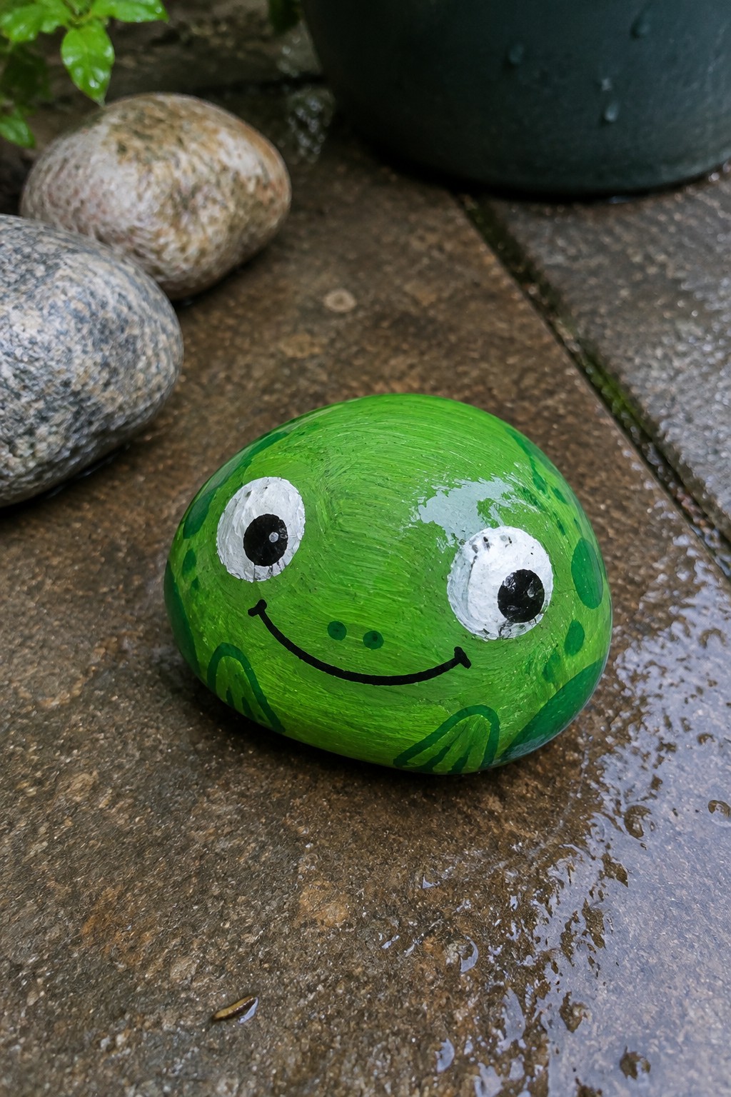 Green frog-painted rock with big white eyes and smile on wet pavement