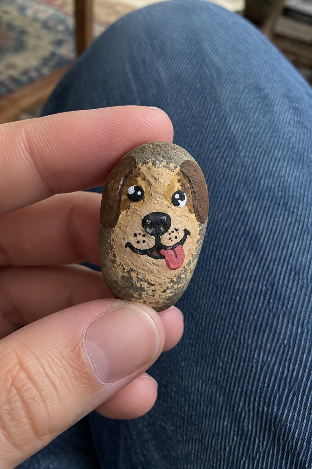 Hand holding oval rock painted as dog with brown ears and pink tongue.